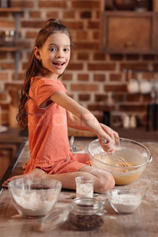 \'side view of girl sitting on table and mixing ingredients for cake, stock photo
