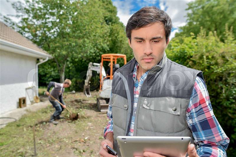Man stood outside looking at tablet, digger in background, stock photo