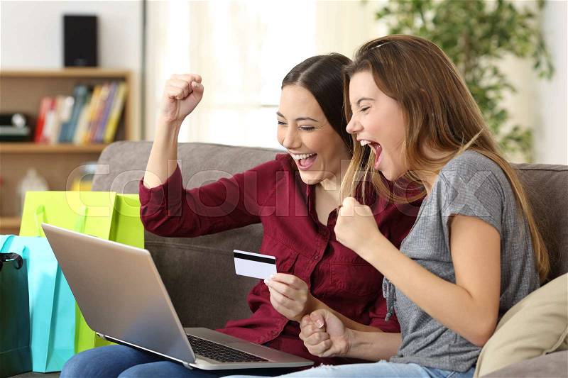 Two excited friends buying online with credit card and a laptop sitting on a sofa in the living room at home with a homey background, stock photo