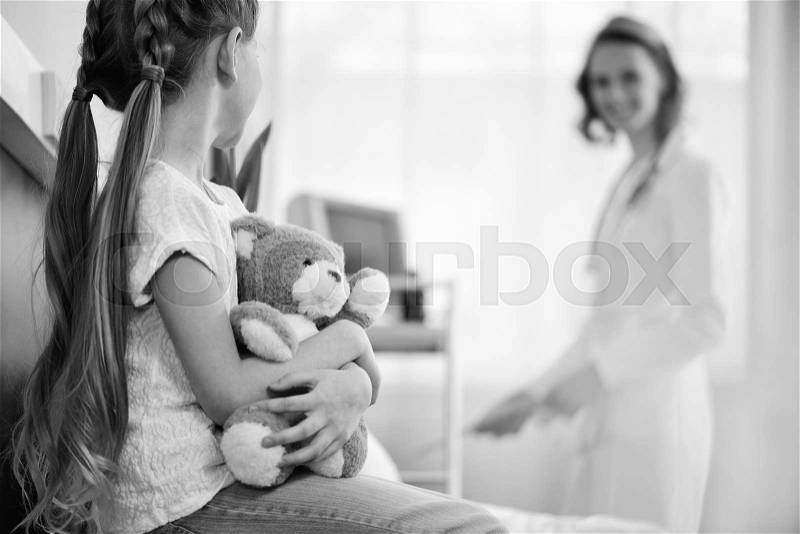 Girl with teddy bear in hospital chamber with doctor behind, black and white photo, stock photo