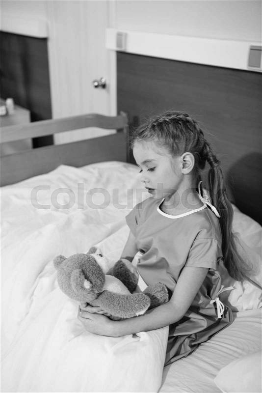Serious little girl with teddy bear sitting on hospital bed, black and white photo, stock photo