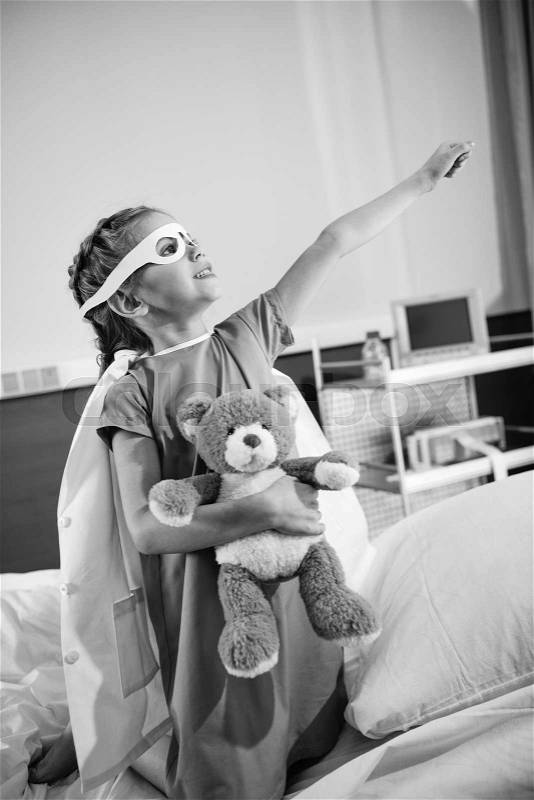 Black and white photo of little girl superhero playing with teddy bear i hospital, stock photo