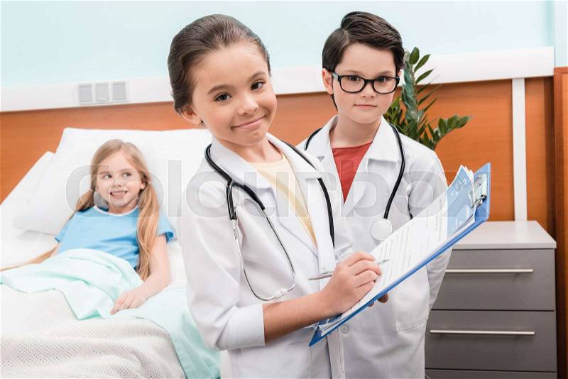 Kids with stethoscopes playing doctors near little patient lying in hospital bed, stock photo