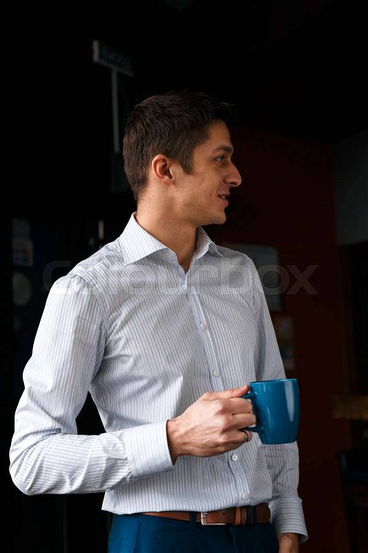 Coffee break. Confident young handsome man in blue pants and white shirt holding cup with hot drink and smiling while standing, stock photo