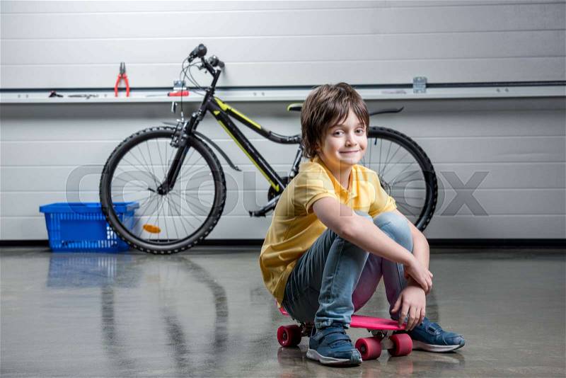 Adorable boy in sitting on penny board and looking at camera, bicycle behind, stock photo