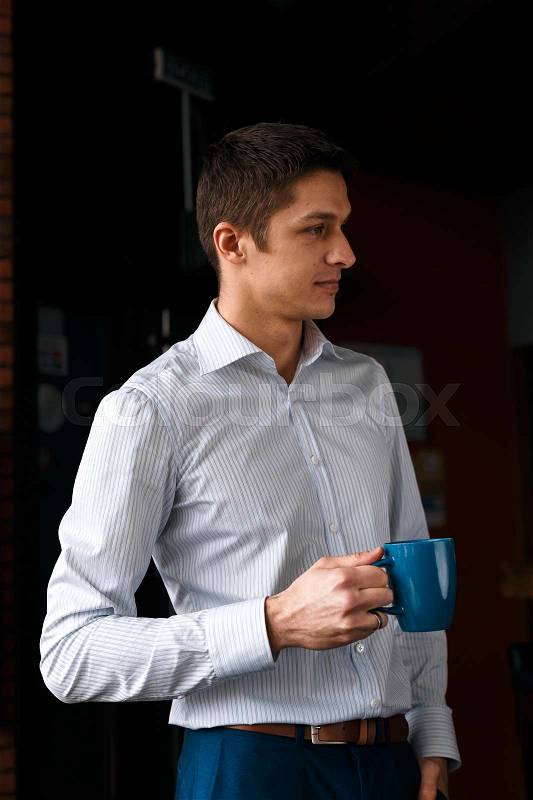 Coffee break. Confident young handsome man in blue pants and white shirt holding cup with hot drink and smiling while standing, stock photo