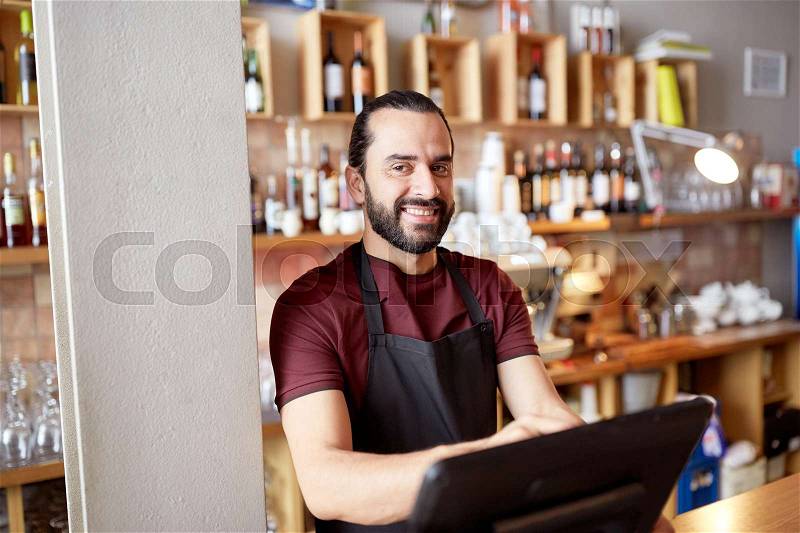 Small business, people and service concept - happy man or waiter in apron at counter with cashbox working at bar or coffee shop, stock photo