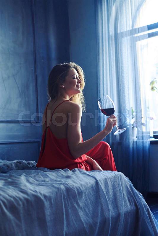 Back view of a young elated woman with glass of red wine sitting on the bed. Woman sitting on the bed in front of the window. Obscure room interior , stock photo