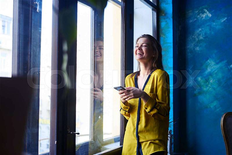 Laughing young girl with telephone in hands standing next to the window. Girl in yellow blouse. Darkblue walls, obscure atmosphere , stock photo