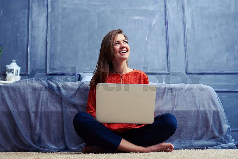 Low angle of a female sitting cross-legged with laptop looking upward. Young creative woman sitting in the floor. Barefoot young girl working at home , stock photo