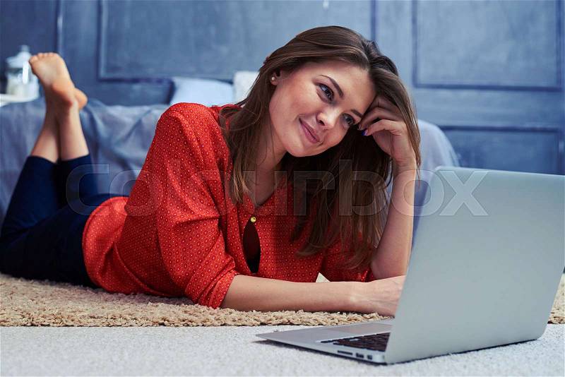 Close-up of an embarrassed Caucasian young woman lying on floor using laptop. Open silver laptop. Girl working at home , stock photo