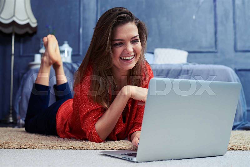 Close-up of smiling young girl lying on the floor in front of laptop. Open silver laptop standing on the floor. Female looking at the screen , stock photo