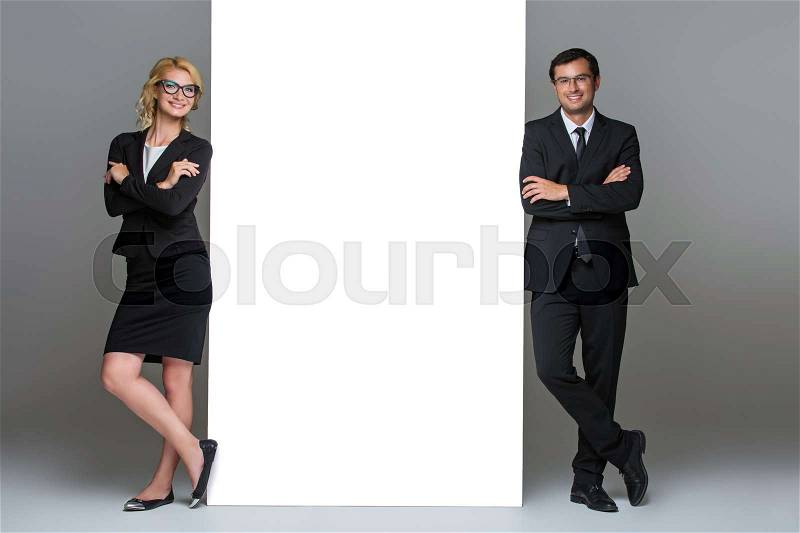 Beautiful business lady and handsome businessman standing near big empty poster. Happy expression. On gery background. Copy space, stock photo