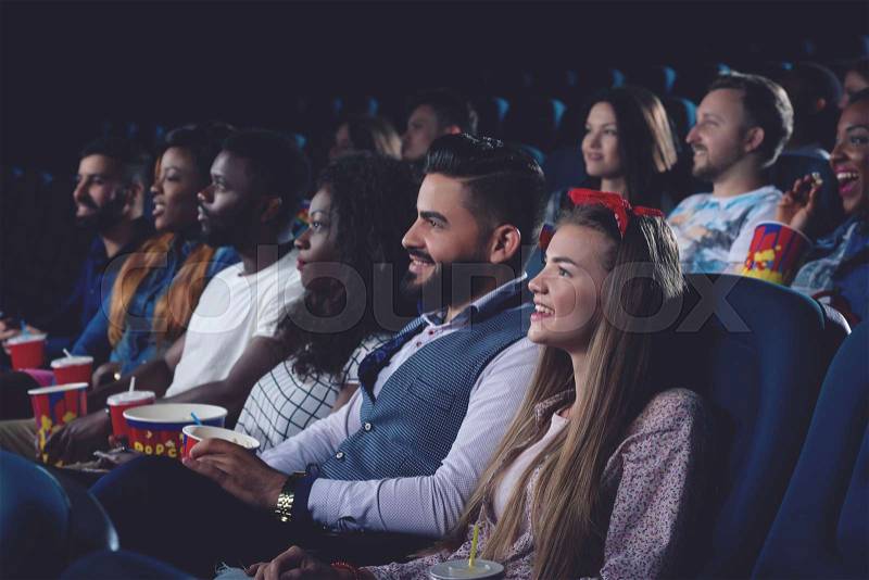Side view of young women and men watching movie in modern cinema hall and smiling. Group of people eating popcorn and spending free time in cinema together, stock photo