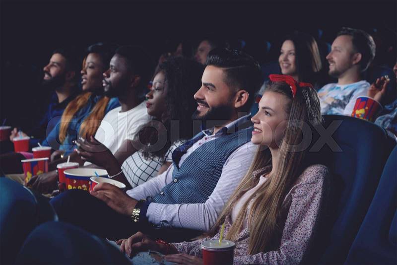 Company of international people watching interesting movie in modern cinema hall. Men and women watching at big projector and emotional smiling, stock photo