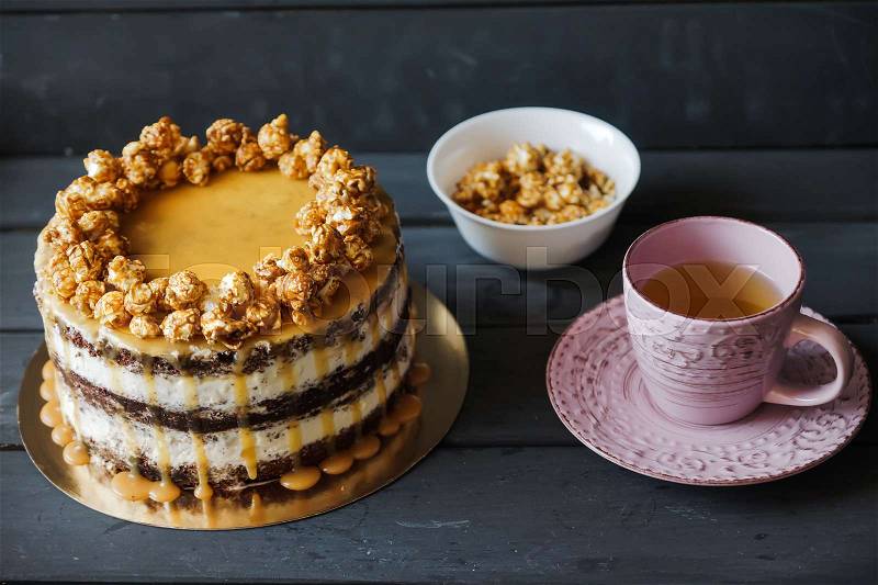 Delicious cake with caramel popcorn and caramel sauce and plate with popcorn and pink cup of tee, stock photo