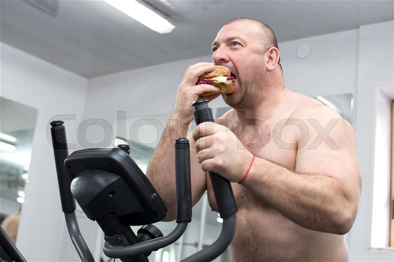A big fat hungry man eats a hamburger with meat and cheese in the gym, stock photo