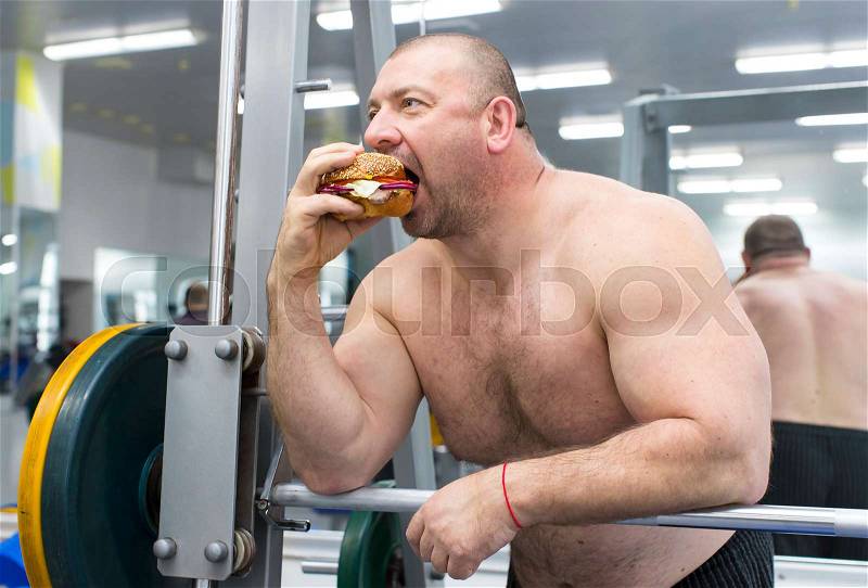 A big fat hungry man eats a hamburger with meat and cheese in the gym, stock photo