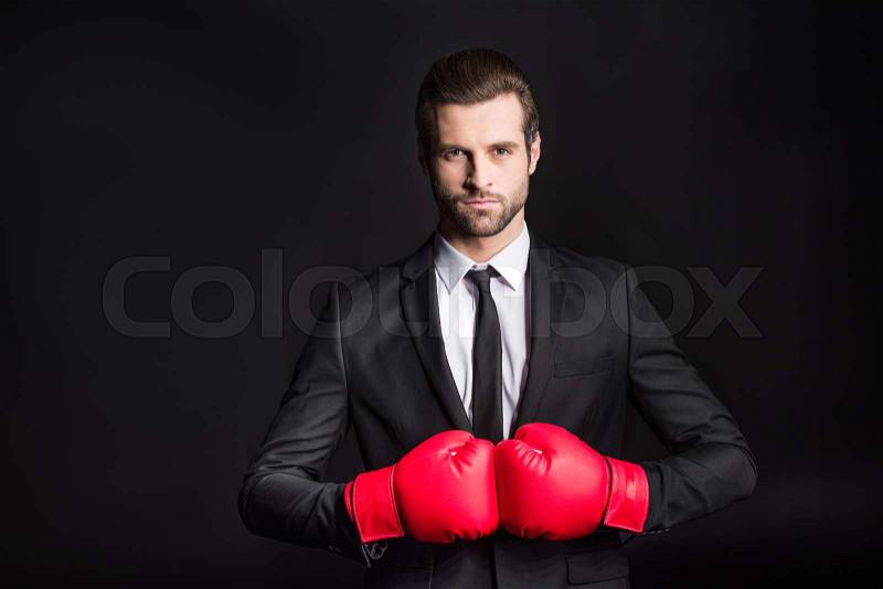 Confident young businessman in red boxing gloves looking at camera on black, stock photo