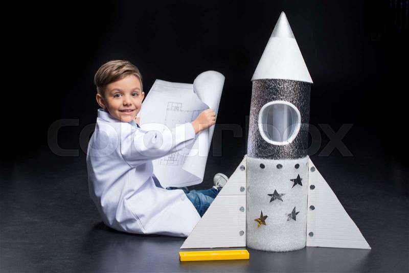 Little boy in white coat holding blueprint while sitting near toy rocket and smiling at camera, stock photo