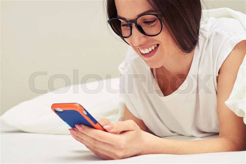 Side view of an elated woman using phone while lying on the bed at home. Woman covered with white blanket, having rest on the bed, stock photo