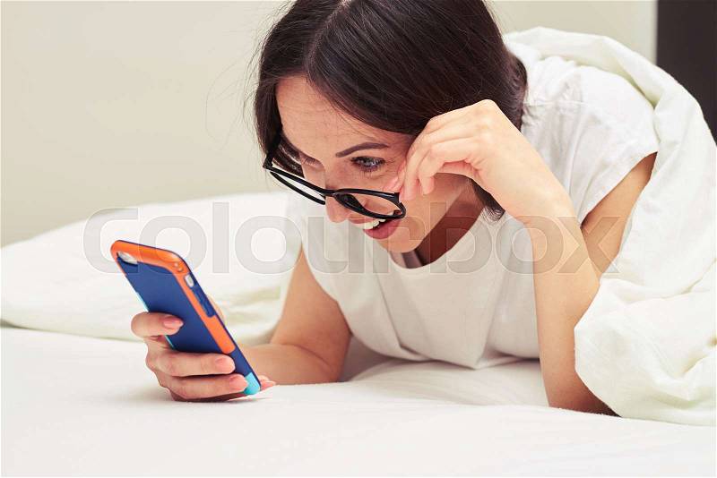 Close-up shot of a surprised woman holding glasses while reading something on the telephone. Impressed female noticed something. Lying on the bed at home, stock photo