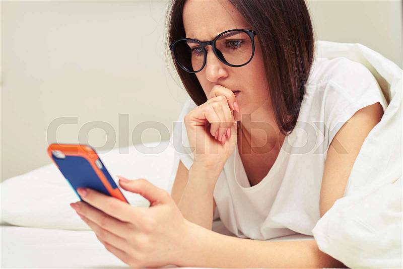 Close-up shot of preoccupied female looking at the telephone while lying on the bed. Female looking attentively at the screen of the telephone , stock photo
