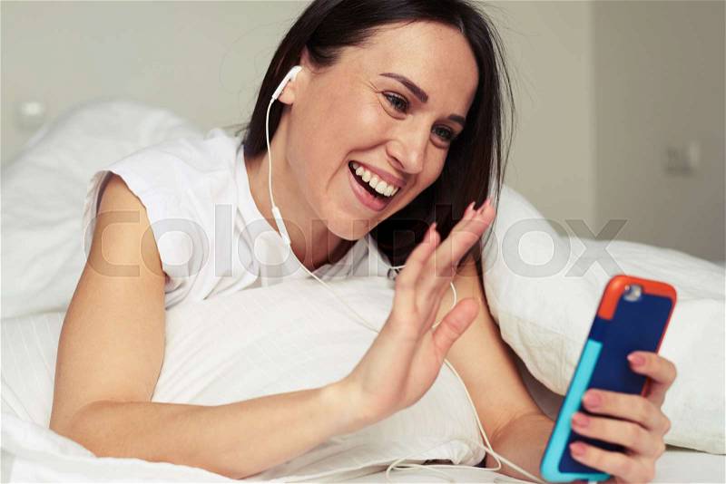 Close-up shot of Caucasian female communicates with someone via smartphone while lying on the bed. Using headphones. Having rest while lounging on the bed at home , stock photo