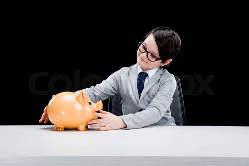 Smiling little boy businessman sitting at table with piggy bank, stock photo