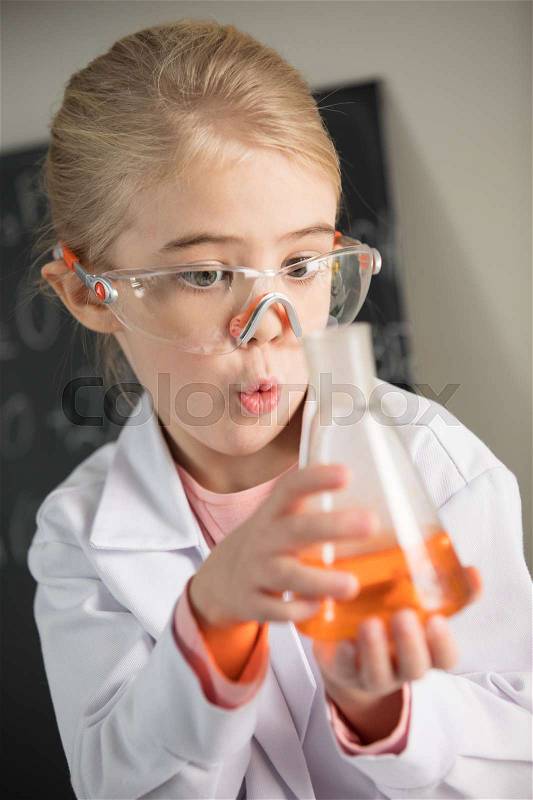 Exited girl wearing lab coat and protective glasses holding flask with orange reagent , stock photo