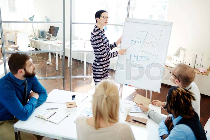 Beautiful middle-aged team leader standing at whiteboard and pointing at diagram of company productivity, multiethnic group of workers listening to her and taking notes, stock photo
