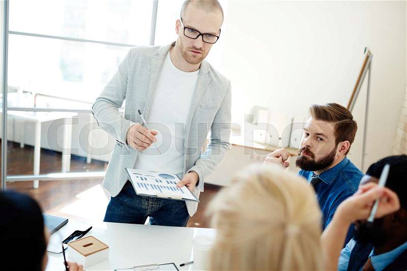 Multiethnic group of managers sharing ideas at meeting, handsome team leader in eyeglasses standing at table and listening to his female colleague with interest, stock photo