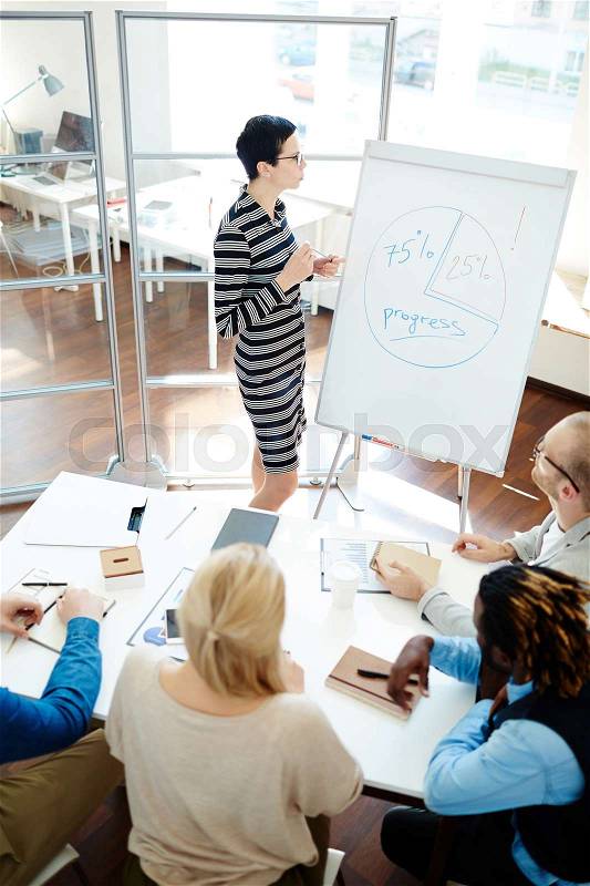 High angle view of pensive businesswoman looking at whiteboard with productivity diagram and thinking over possible ways of increasing company performance, her colleagues sharing ideas, stock photo