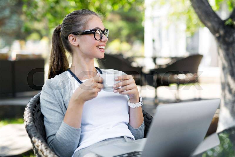 Cheerful young employee in stylish eyeglasses looking away while drinking tea in outdoor cafe, modern laptop standing on table, stock photo