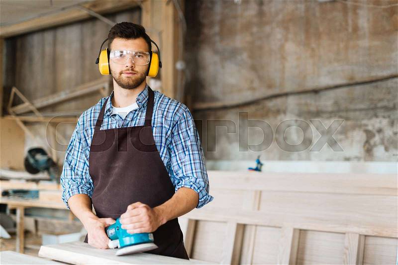 Portrait of bearded craftsman with electric sander in hands distracted from work in order to pose for photography while standing at wooden timber, stock photo
