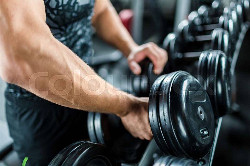 Closeup shot of muscular male arm picking up heavy dumbbell weights from equipment rack in modern gym, stock photo