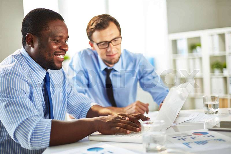Collaborative work on new computer application: Afro-American programmer writing code on laptop while his superior giving guidance, stock photo