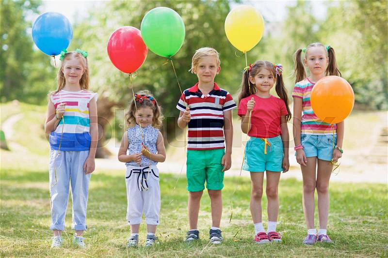 Happy children in summer wear standing in line on green lawn, holding colorful balloons and looking at camera, stock photo