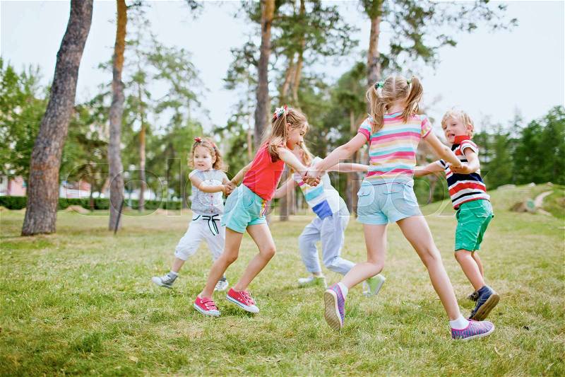 Group of children holding hands and dancing in circle on green lawn in park on beautiful summer day, stock photo