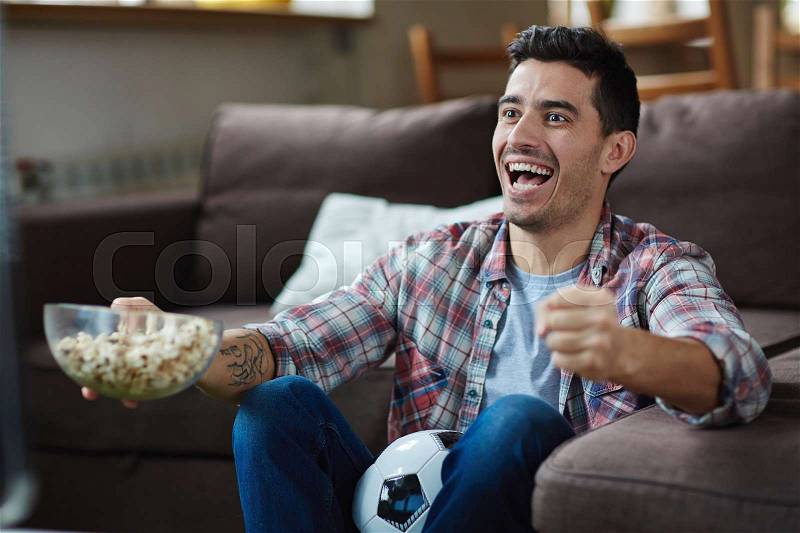 Portrait of excited man watching sports match on TV and cheering joyfully while eating popcorn, stock photo