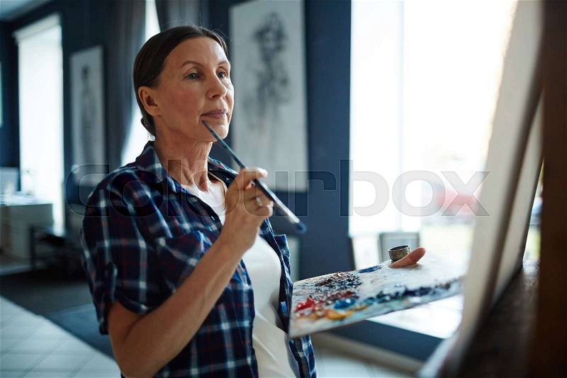 Pensive female with paintbrush and palette looking at her picture on easel, stock photo