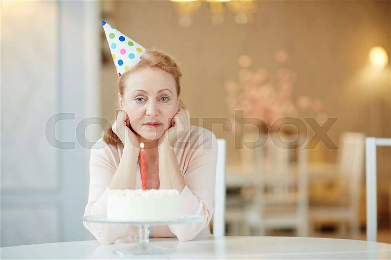 Portrait of sad lonely mature woman sitting alone at birthday table with cake , wearing party hat and looking at camera, stock photo