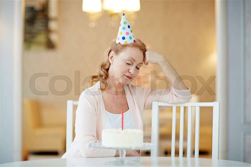 Portrait of sad lonely mature woman sitting alone at birthday table with cake, wearing party hat and waiting for guests, stock photo