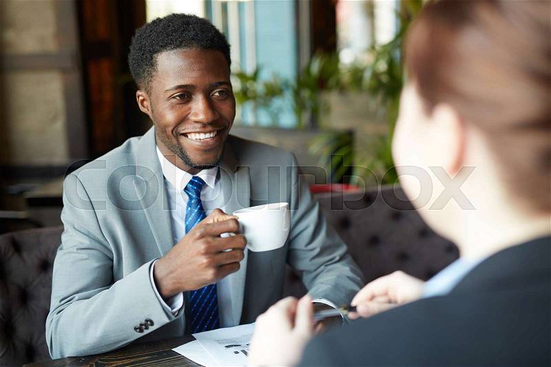 Two business people meeting in modern cafe: African-American man wearing business suit smiling while looking at his partner and drinking coffee, stock photo