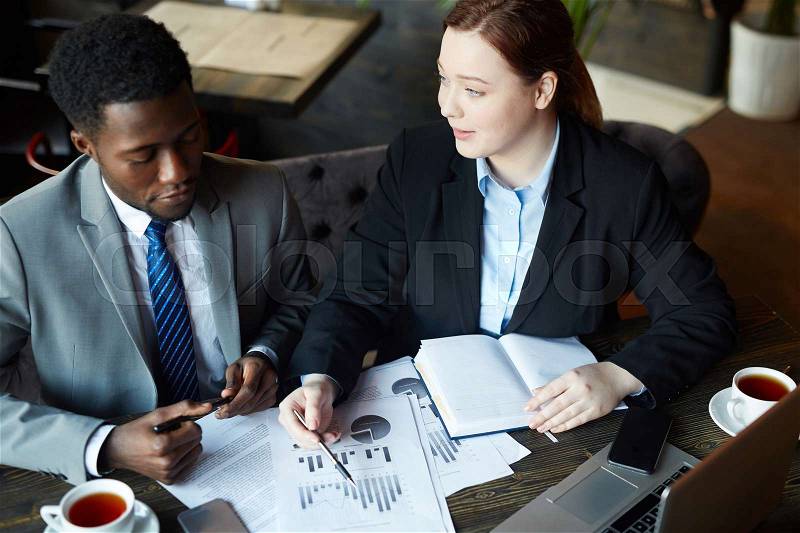 Portrait of two business people meeting in modern cafe: Young professional woman pointing to statistics graphs while explaining data to African –American colleague, stock photo