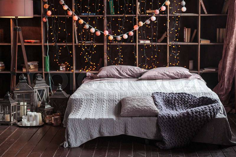 Coziness, comfort, interior and holidays concept - cozy bedroom with bed and garland lights at home. A rack with books behind the bed. Candles, a lamp and a lamp stand near the bed. Plaid hand-knitted, stock photo