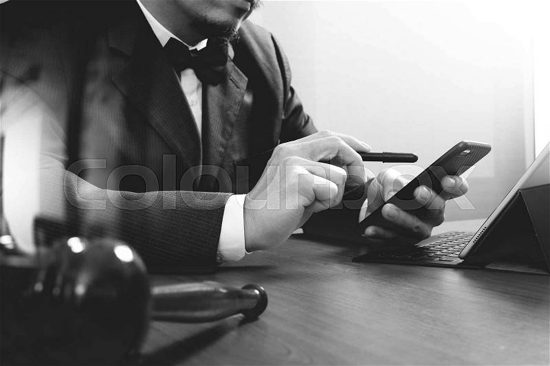Justice and Law context.Male lawyer hand working with smart phone,digital tablet computer docking keyboard with gavel and document on wood table,black and white, stock photo