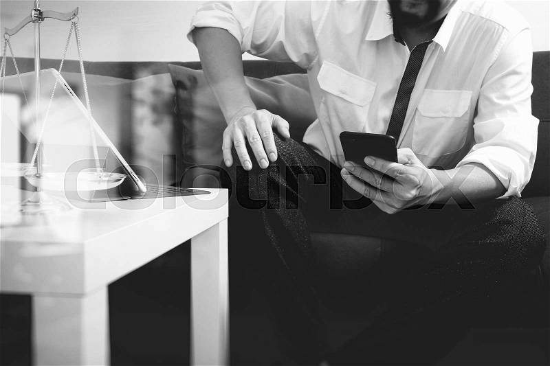 Justice and Law context.Male lawyer hand sitting on sofa and working with smart phone,digital tablet computer docking keyboard with gavel and document on living table at home,black white, stock photo