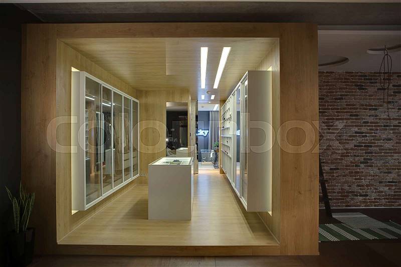 Stylish wooden walk-in closet with white wardrobes, shelves, stand and mirrors. There are luminous lamps. On the right side there is a brick wall and small carpets on the floor. Horizontal, stock photo