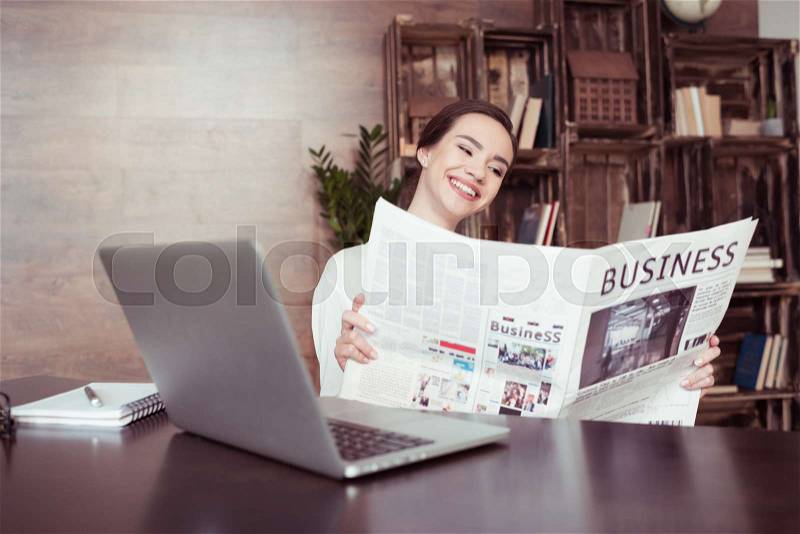 Young smiling businesswoman reading newspaper at desk with laptop, stock photo